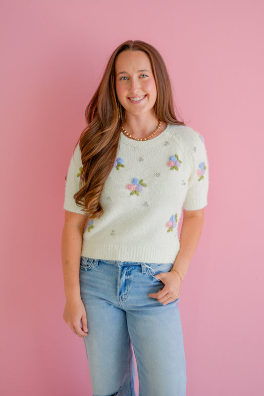 In February Bloom and Sparkle Sweater featuring a pastel floral pattern and cozy knit fabric, displayed on a hanger.